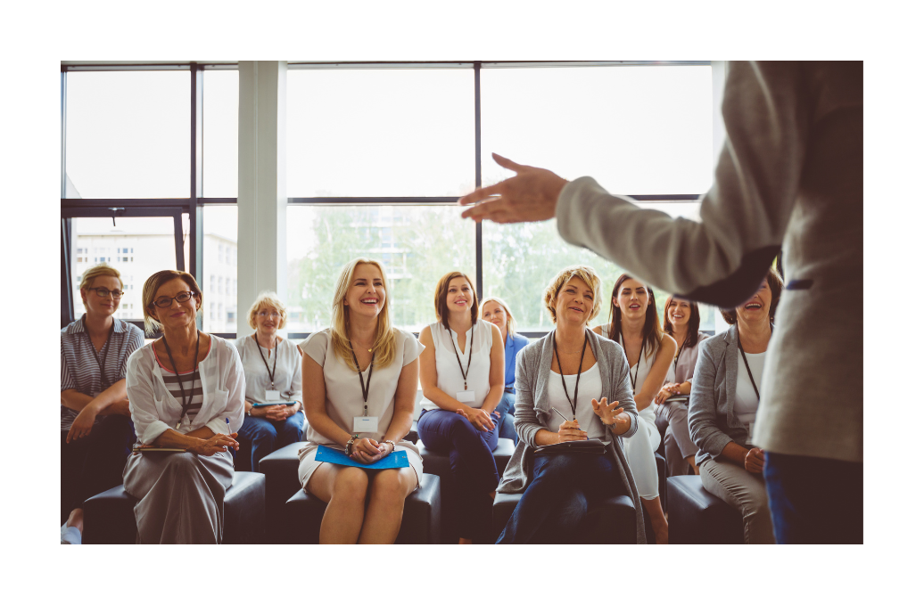 A group of women seated, smiling, and listening attentively to a speaker in a bright, modern conference room.