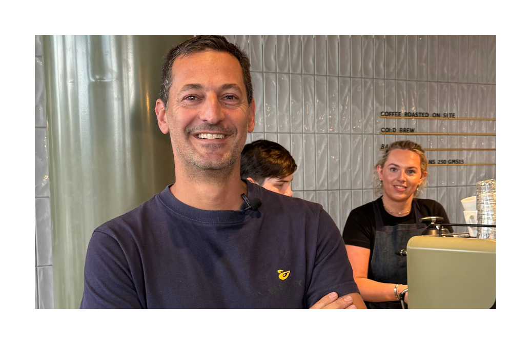 A man smiling confidently with arms crossed in front of a coffee shop counter, with a barista working in the background.