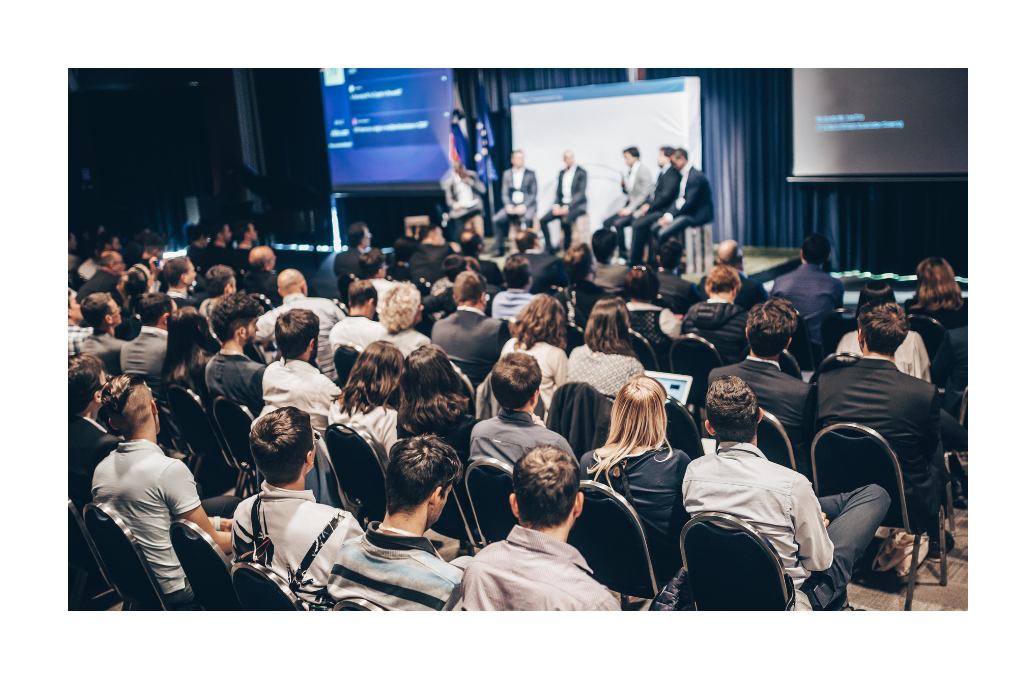 A large audience attending a panel discussion in a dimly lit conference room with speakers on stage in the background.