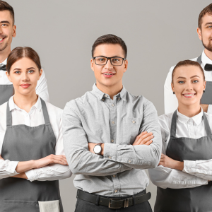 A group of five hospitality workers standing confidently, wearing uniforms with aprons and bow ties, with one person in the center wearing a button-up shirt and glasses, all smiling at the camera.