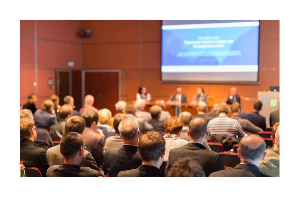 An audience listening to a panel discussion in a well-lit conference room, with the focus on a speaker at the front.