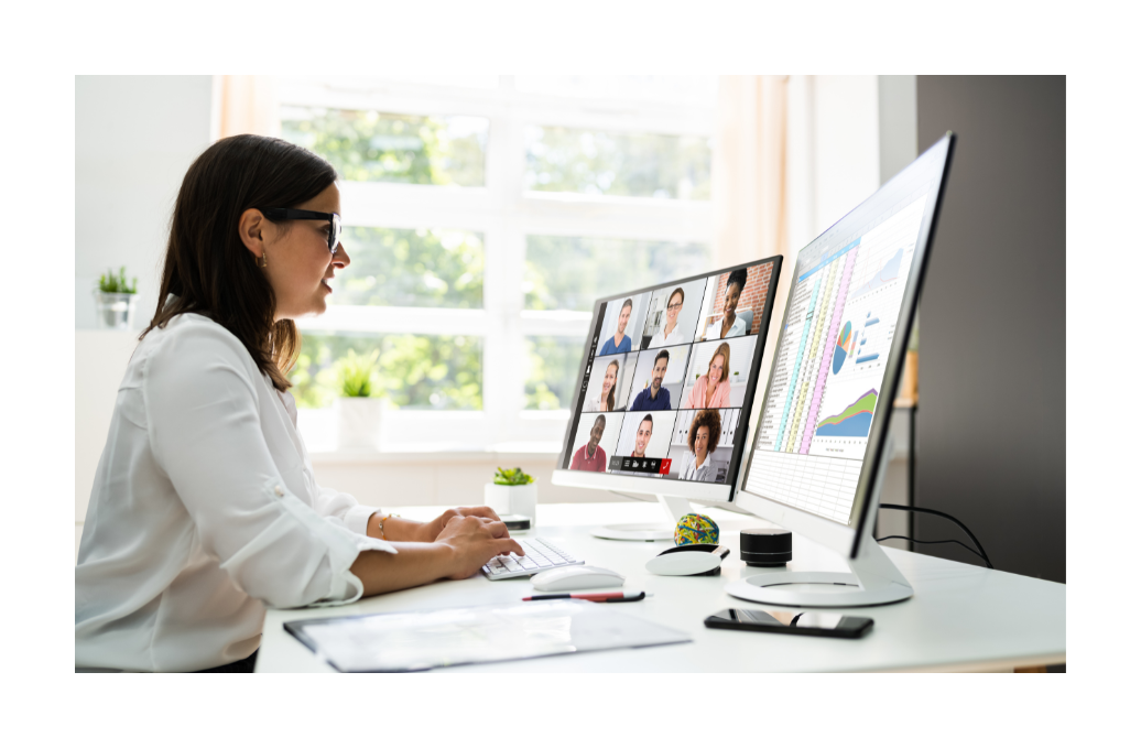 A woman in a white shirt participating in a virtual meeting, looking at a computer monitor with multiple people in a video call.