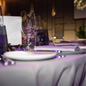 A close-up shot of a formal dining table set with white plates, utensils, and wine glasses. The table is draped with a light-coloured tablecloth, and the setting is softly lit with a purple hue.