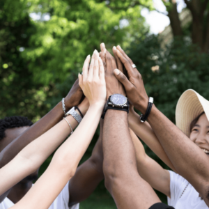 A diverse group of people outdoors, raising their hands together in a high-five gesture, symbolizing teamwork, unity, and collaboration. The background features greenery, indicating a natural, outdoor setting.