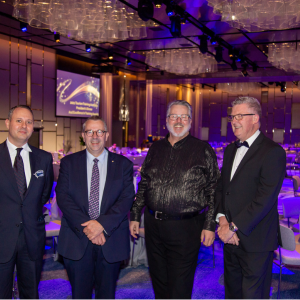 Four men in formal attire, including suits and tuxedos, are posing together in a large, well-decorated event hall with purple lighting.