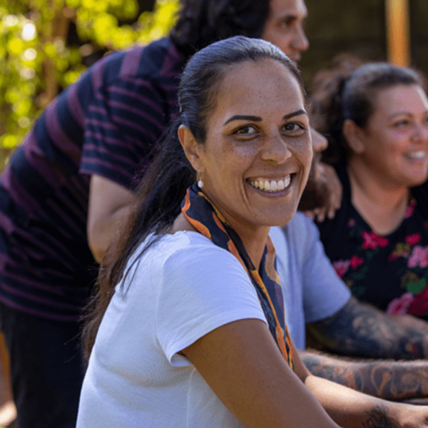 A woman sitting outside with a group of people, smiling warmly at the camera during a community gathering or casual event.