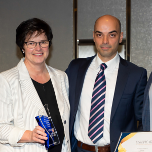 A woman in a light-coloured blazer is holding a blue trophy, standing next to a man in a suit and tie who is holding a framed certificate. They are posing together in a formal setting.