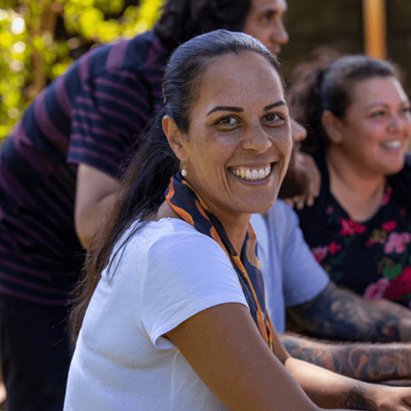 A woman sitting outside with a group of people, smiling warmly at the camera during a community gathering or casual event.