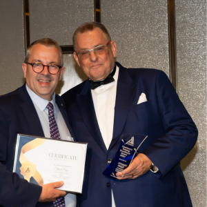 Two men in formal attire are posing for a photo. The man on the left is holding a framed certificate, while the man on the right is holding a blue trophy. Both are smiling.