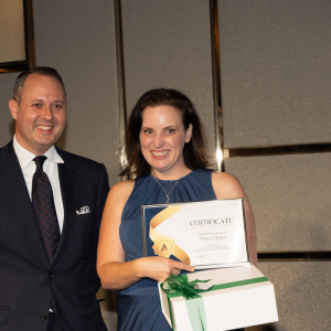 A man in a dark suit is standing next to a woman in a blue dress who is holding a certificate and a gift box with a green ribbon. They are smiling and posing in a formal setting.