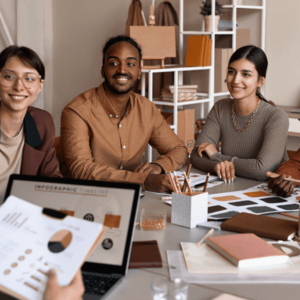 A diverse group of colleagues sitting around a table in a meeting room, discussing design plans. A laptop is open with a presentation on the screen, and various materials are spread out on the table.