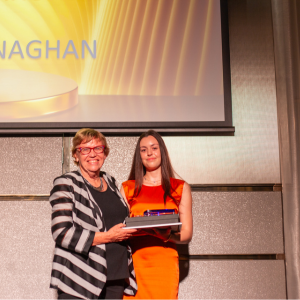 Two women are standing on stage in a formal setting. One is wearing a striped blazer, and the other is in a bright orange dress. They are holding a certificate and a small award.