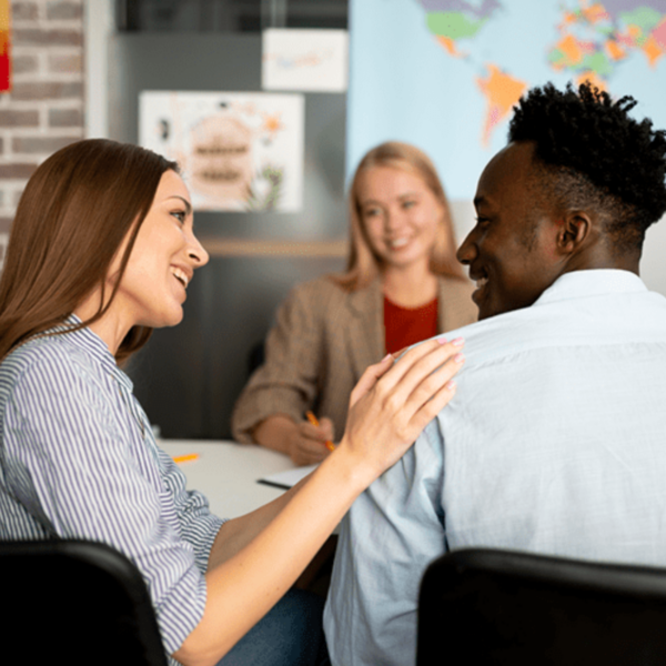 A woman smiling and placing her hand on a man's shoulder, engaging in a friendly conversation. Another woman in the background is watching them and smiling. The setting is a casual office or meeting room.