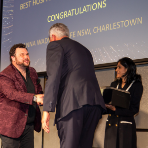 A man in a red velvet blazer is shaking hands with another man in a dark suit on a stage. A woman in a black and white outfit stands nearby holding a box, while a congratulatory message is displayed on the screen behind them.