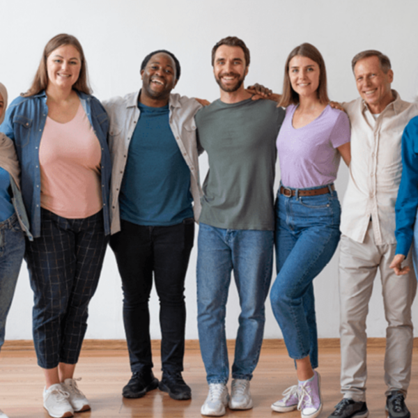 A group of seven people standing together, arms around each other, smiling for the camera. They are casually dressed and standing in front of a plain wall.