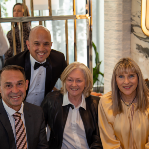 A group of four people posing together in a well-lit, elegant setting. Two women are seated in the front, with one wearing a black leather jacket and the other in a gold satin blouse. Two men are standing behind them, one in a black suit and bow tie, and the other in a dark striped suit and tie.