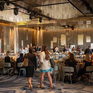 A wide-angle view of a formal event in a spacious, modern venue with reflective, geometric wall decorations. People are mingling around tables set with drinks and snacks, and a few individuals are walking through the space.