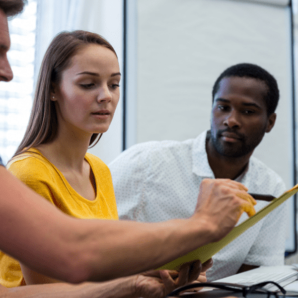 hree colleagues, two men and one woman, engaged in a discussion, with the man in the foreground explaining something using a document.