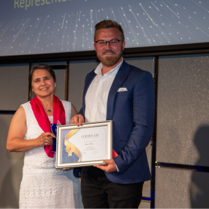 A man in a blue blazer and white shirt holding a framed certificate poses with a woman in a white dress who is holding a small trophy. They are standing on a stage with a screen behind them displaying text.