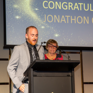 A man in a light gray suit and black shirt is speaking at a podium at the TTA Training awards, with another woman standing behind him. A large screen in the background displays congratulatory text.