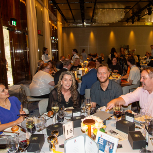 A lively dining scene at a formal event. Several people are seated around a table, engaged in conversation and enjoying food and drinks. The table is set with wine glasses, small boxes, and table decorations.