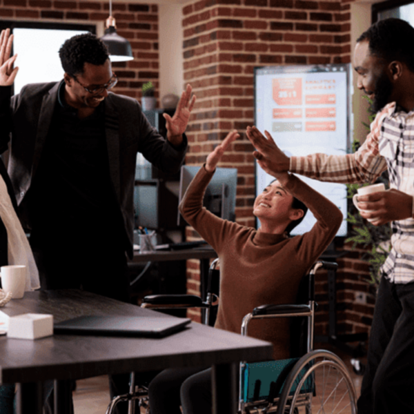 A joyful scene in an office setting where two men are giving high-fives to a young woman in a wheelchair. The atmosphere is celebratory, with smiles and raised hands all around.