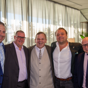 A group of five men dressed in business attire are standing together, posing for a photo. They are smiling and appear to be in a bright, indoor setting with curtains and plants in the background.