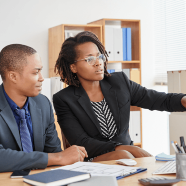 Two professionals in business attire, a man and a woman, are sitting at a desk. The woman is pointing at a computer screen, explaining something to the man, who is listening intently.