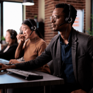 Call center operators wearing headsets are working at their desks in a modern office. They are focused on their tasks, with one person in the foreground and others in the background.