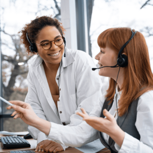 Two women working as call center operators are smiling and talking to each other. Both are wearing headsets and sitting at a desk in a bright office with large windows.