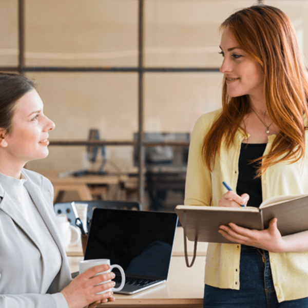 Two women are engaged in a conversation in an office. One is holding a notepad and pen, while the other is holding a coffee mug. They are smiling and appear to be discussing something in a friendly manner.