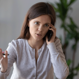 A woman with long dark hair, wearing a light gray shirt, is speaking on the phone. She looks concerned or focused, gesturing with her hand as she talks.