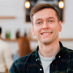 A young man in a checkered shirt is smiling at the camera in a casual, well-lit setting, possibly a café or informal workspace, with blurred people in the background.