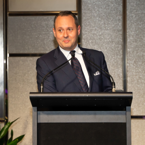 A man in a dark suit and patterned tie is standing at a podium with a microphone, appearing to speak or present. The background features a modern, metallic wall design.