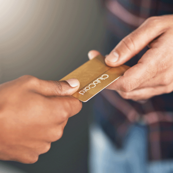 A close-up of two hands exchanging a gold-coloured card labeled "Clubcard." The focus is on the hands and the card, with a blurred background.