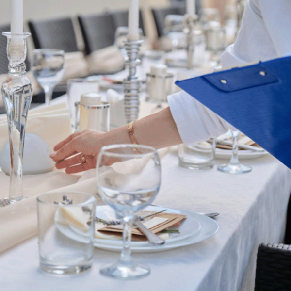 A person is setting a formal dining table, adjusting the placement of a napkin. The table is elegantly set with glassware, candles, and white linens.