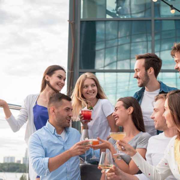 A group of young adults socialising outdoors, holding drinks, and smiling. The setting is a rooftop or patio area with a modern building in the background.