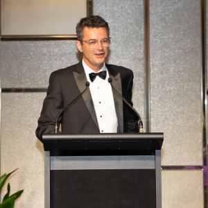 A man in a black tuxedo with a bow tie is speaking at a podium with a microphone. The background is a sleek, modern wall with a soft glow.