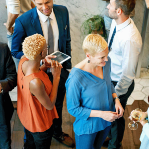 A diverse group of professionals mingling and chatting at a networking event. One woman is holding a tablet, and others have drinks in hand. The setting is informal with a lively atmosphere.