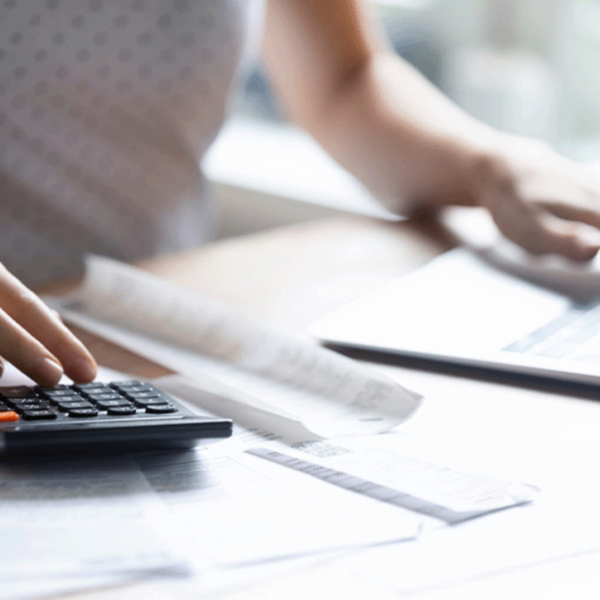 A person using a calculator with one hand while other paperwork and a laptop are visible on the desk. The scene suggests a focus on financial tasks or budgeting.