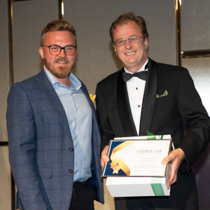 Two men are posing together at the President's Dinner & Training Awards, one in a blue blazer and the other in a black tuxedo. The man in the tuxedo is holding a certificate and a white box with a green ribbon. Both are smiling.