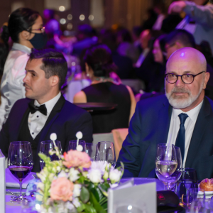 Two men in formal attire at the President's Dinner & Training Awards are seated at a table set with wine glasses and floral arrangements. The background is filled with other guests at a formal event, with dim lighting creating a purple ambiance.