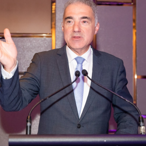 A man in a dark suit and light blue tie is speaking at a podium with two microphones at the President's Dinner & Training Awards. He is gesturing with one hand, and the background features a modern, well-lit wall.