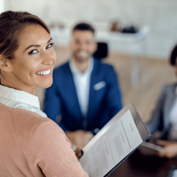 A woman holding her resume and smiling at the camera with two interviewers blurred in the background.