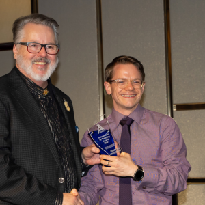 Two men are posing for a photo at the President's Dinner & Training Awards; one is wearing a dark patterned suit and the other a light purple shirt. The man in the purple shirt is holding a blue trophy and smiling.