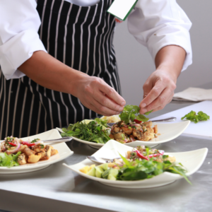 A chef wearing a black and white striped apron is preparing several plates of food, carefully adding fresh herbs to each dish in a professional kitchen setting.