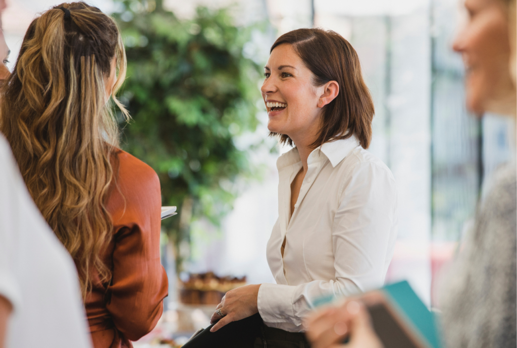 A woman in a white blouse smiling and engaging in conversation with others during a social or professional event.