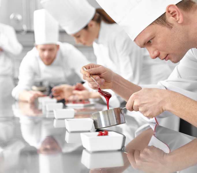 A group of chefs in a professional kitchen, with one chef in the foreground focused on delicately pouring sauce into a dish.