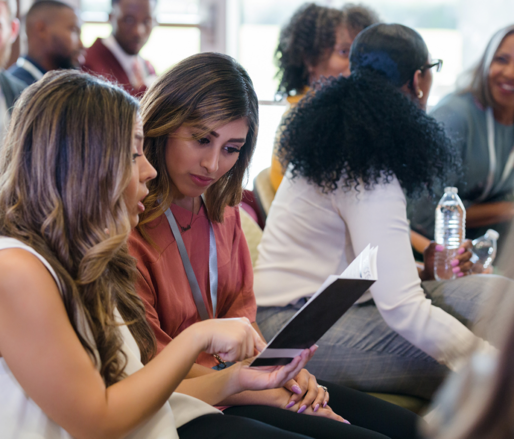 Two women sitting together at an event, engaging in conversation while looking at a pamphlet or program. They are surrounded by other attendees, some of whom are chatting and holding bottled water. The setting appears to be a conference or seminar, with a diverse group of participants in the background.