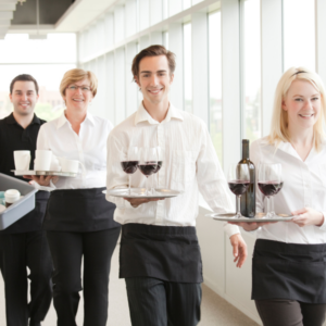 A group of four smiling hospitality staff members are walking down a bright hallway. They are dressed in white shirts and black aprons. The staff members are carrying trays with wine glasses, a wine bottle, and coffee cups, showcasing a professional and welcoming service environment.